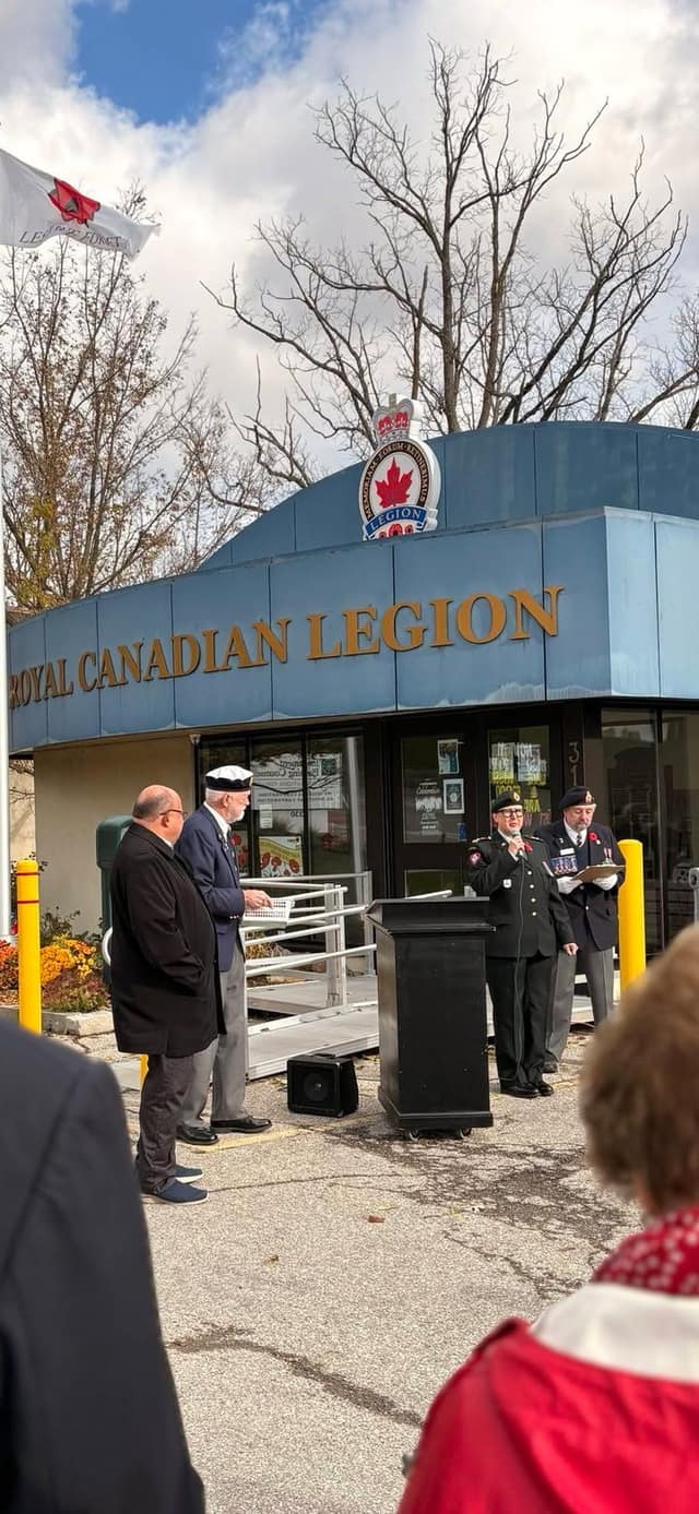 Speakers stand near a podium outside the Royal Canadian Legion entrance during the ceremony, with the poppy flag flying nearby.
