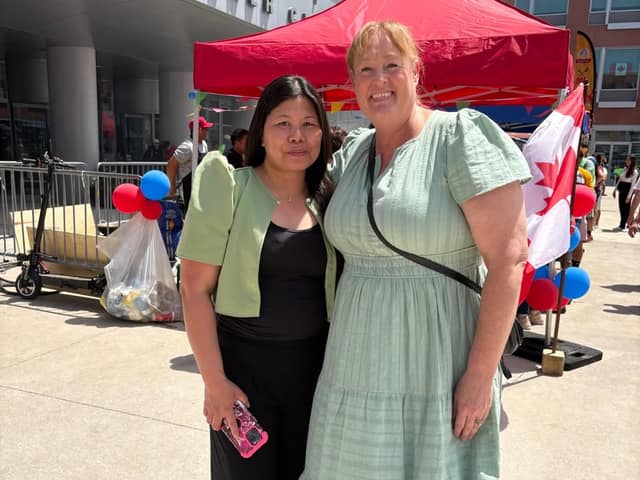 A woman in a green dress poses with a woman in a green jacket holding a phone, beside a Canadian flag and red and blue balloons.