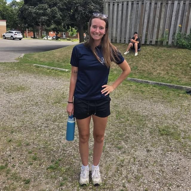 A smiling young woman in a navy camp shirt and black shorts holds a blue water bottle on a gravel lot, with a fence and parked cars behind.