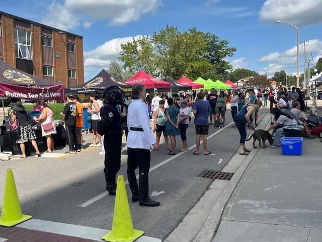 A crowd walks along a closed street past market tents, with two costumed figures in black and white uniforms in the foreground.