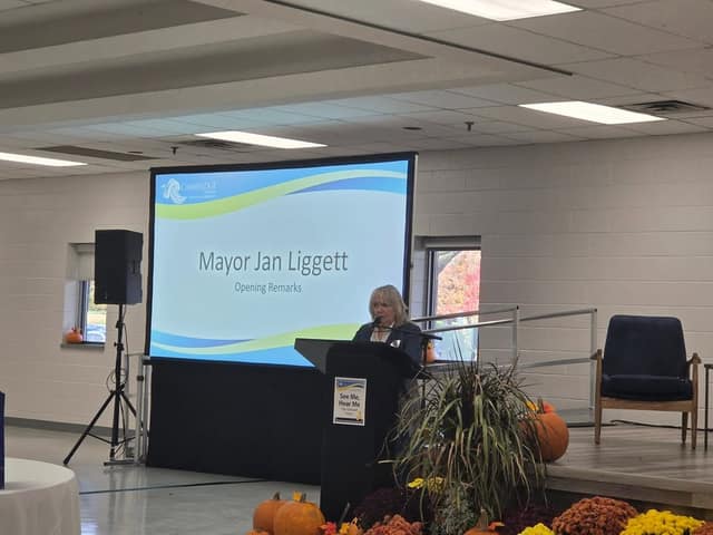 A woman speaks at a podium beside a screen reading "Mayor Jan Liggett, Opening Remarks", with pumpkins and mums in the foreground.