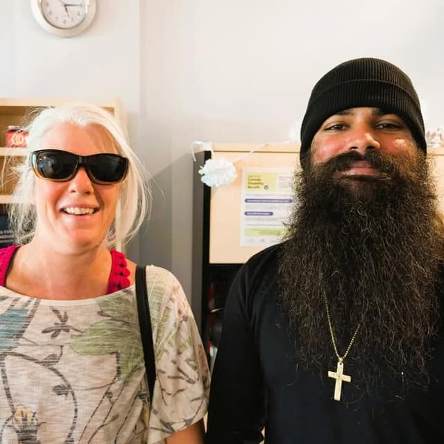 A smiling woman in sunglasses stands beside a man in a black beanie with a long beard and a cross necklace, inside an office.
