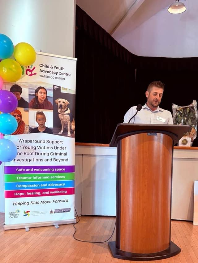 A man speaks at a wooden podium beside a Child & Youth Advocacy Centre banner and balloons, with a bouquet behind him.