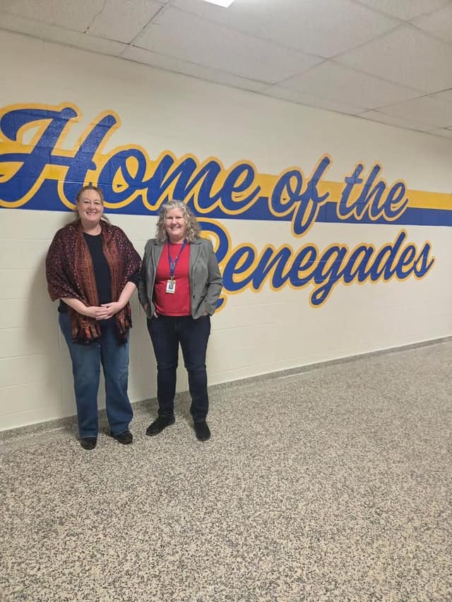 Two women stand in a school hallway in front of a painted wall mural reading "Home of the Renegades."