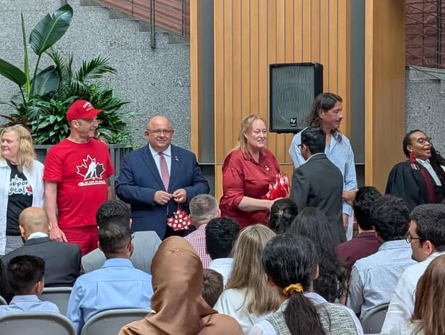 Smiling officials in red and navy stand on a stage holding small paper maple leaves, facing a seated audience at a citizenship ceremony.