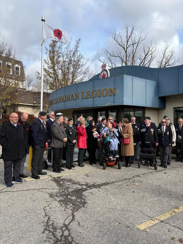 A crowd of veterans, cadets and residents gathers outside the Royal Canadian Legion building beneath a flag with a red poppy.