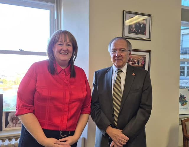 A woman in a red blouse and Senator Jim Quinn in a gray suit pose side by side in an office, with a window and a Kitchener poster behind.