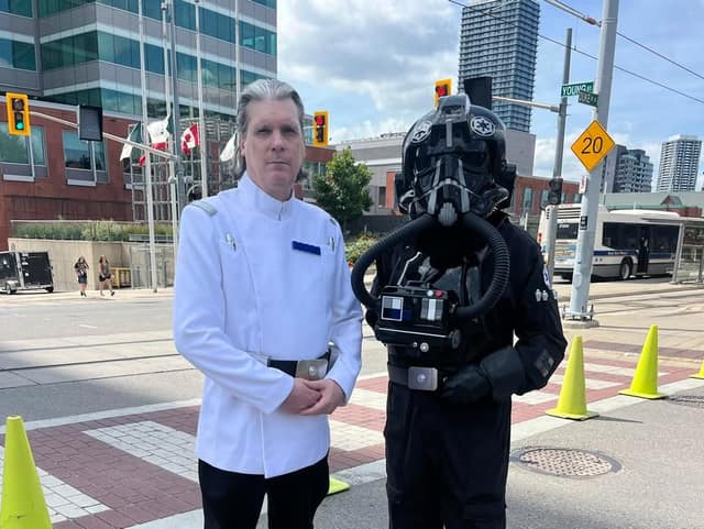 A man in a white uniform jacket stands beside a person in a black helmet and flight suit at a city intersection with a bus behind.