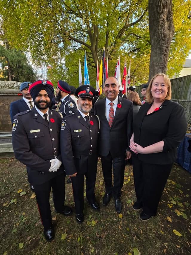 A woman and a man in a suit pose with two uniformed police officers, one in a turban, in front of flags and fall trees.