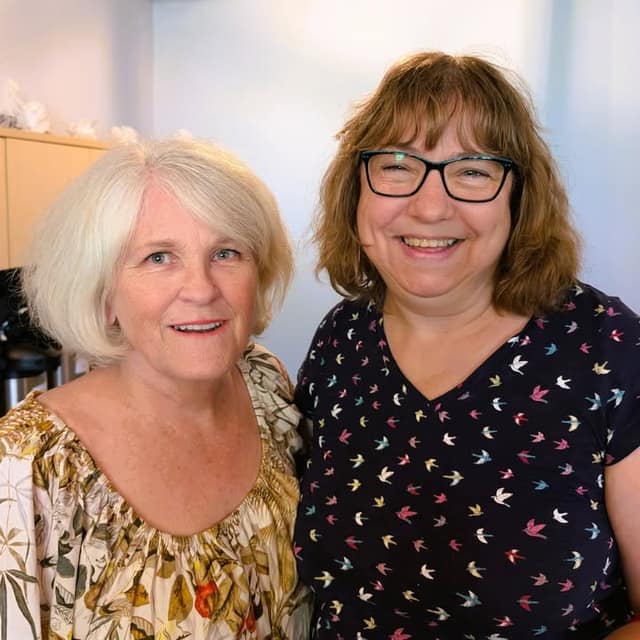 Two smiling women stand close together indoors, one with short white hair in a floral top, the other with glasses in a bird-print shirt.