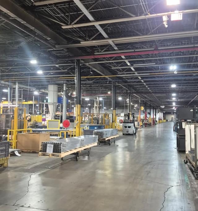 A wide factory interior with high steel ceilings, a forklift in the distance, and carts of metal parts on a concrete floor.