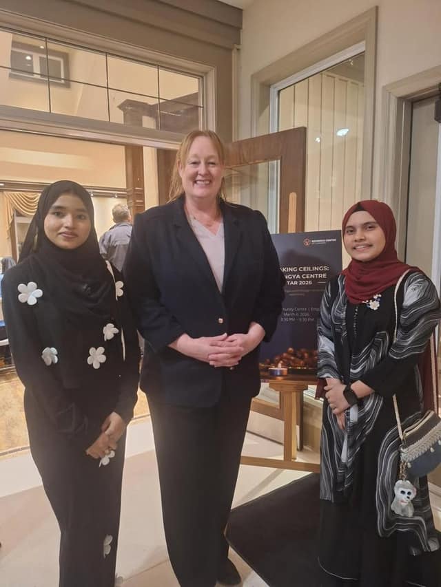 Kelly DeRidder stands between two young women in dark clothing beside an event sign for a Rohingya Centre Iftar dated March 6.