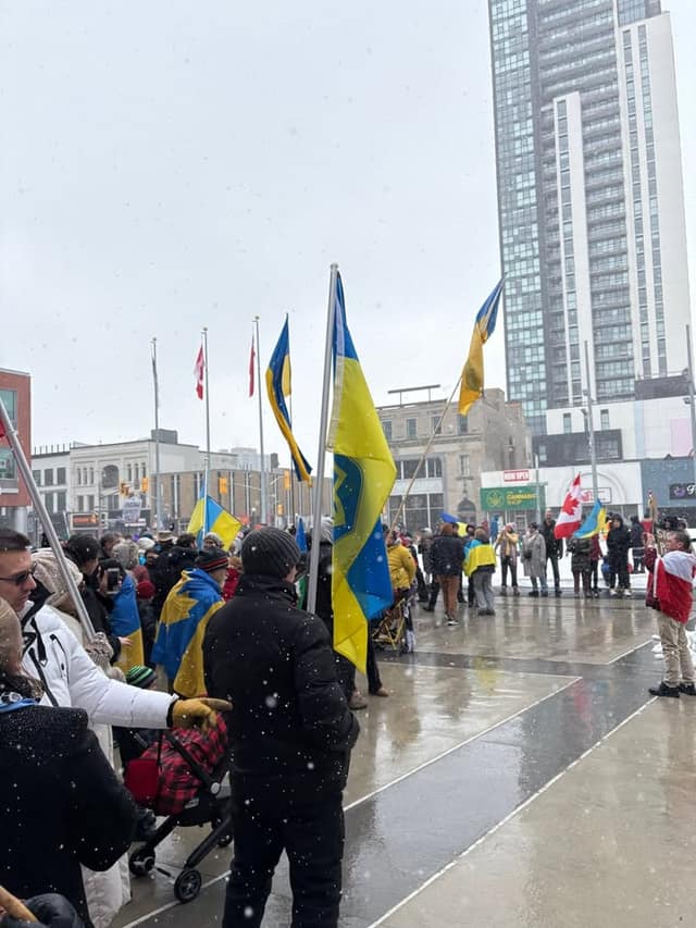 Attendees with Ukrainian and Canadian flags fill a wet city square in light snow, with a tall glass tower in the background.