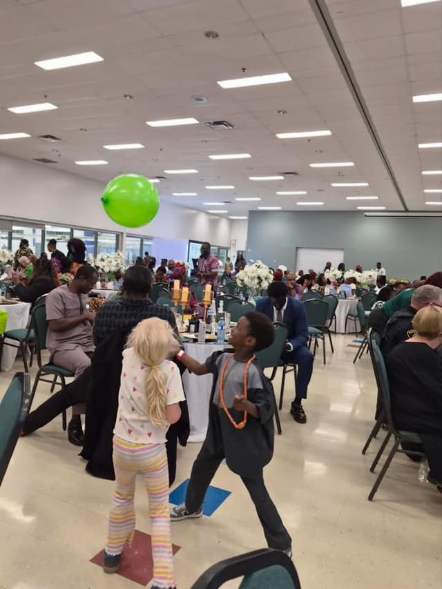 Two children play with a green balloon in a banquet hall, with guests seated at round tables with white flower centrepieces behind them.