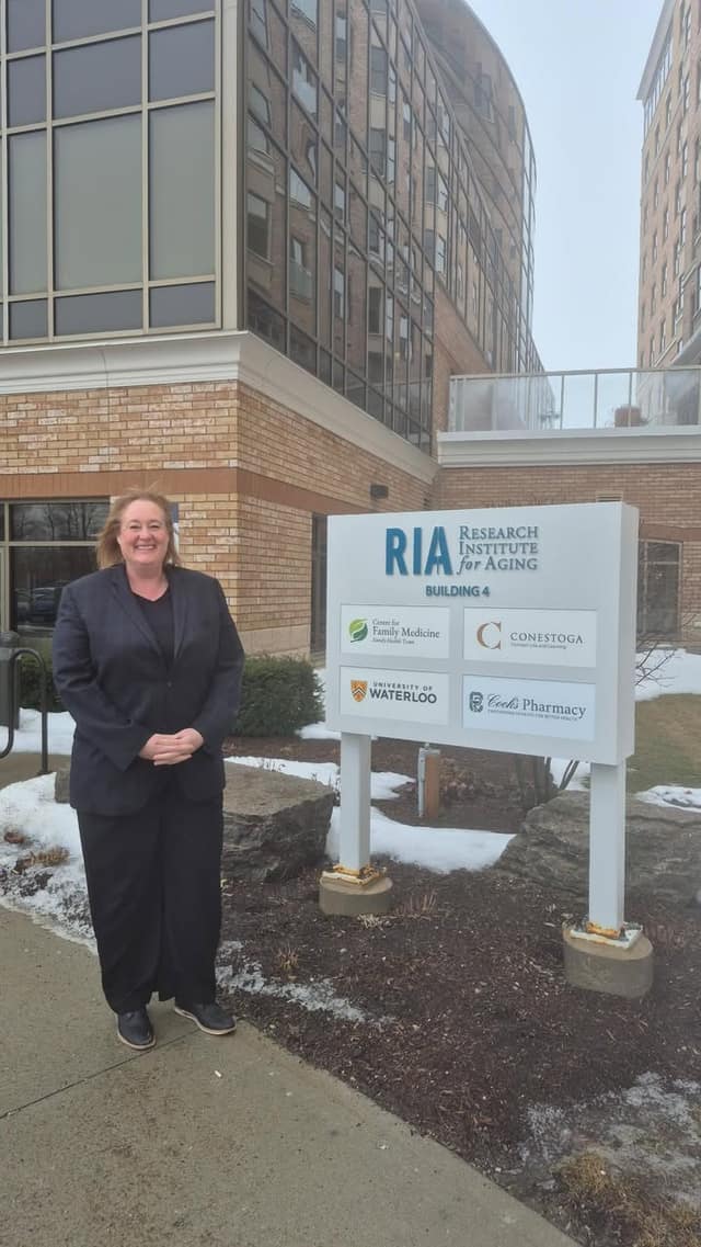 A woman in a dark suit smiles beside a sign reading RIA Research Institute for Aging, Building 4, outside a brick building.
