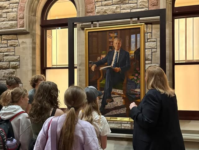 Students and a woman in a dark blazer look at a large gold-framed portrait of a seated man in a suit.
