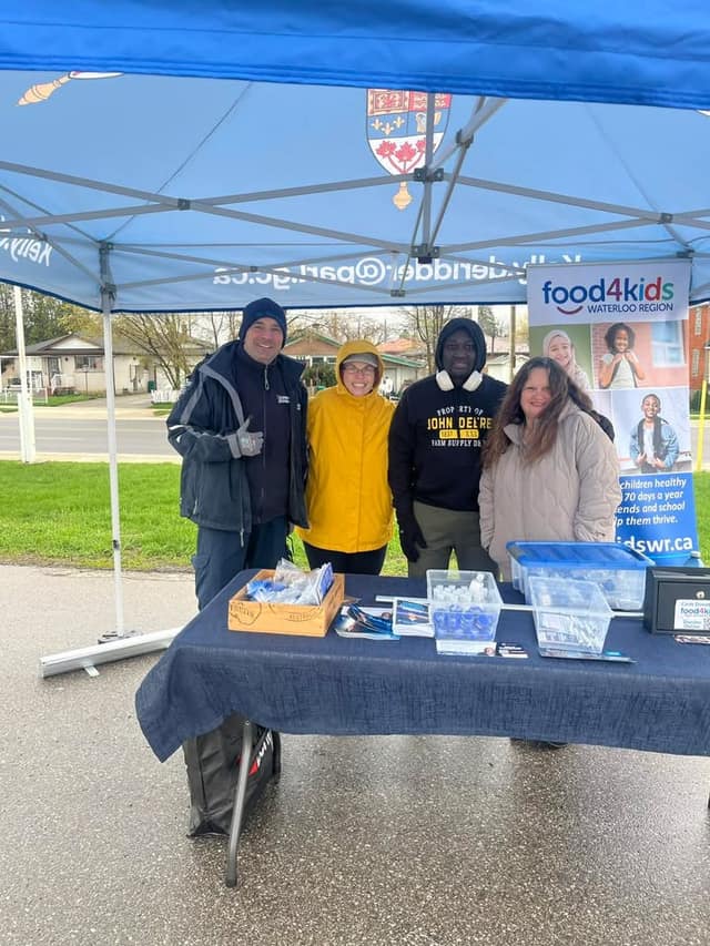 Four volunteers stand smiling behind a table under a blue tent with a Food4Kids Waterloo Region banner on a rainy day.
