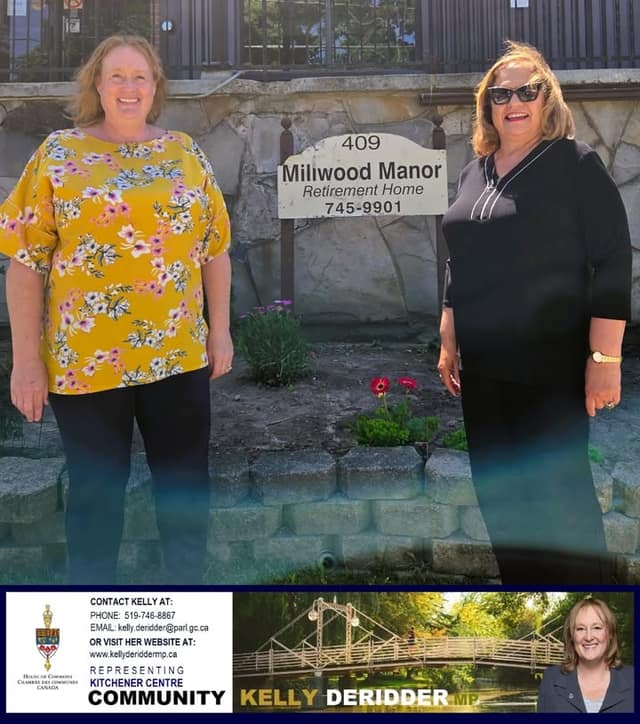 Two women stand in front of a stone wall and a sign reading Milwood Manor Retirement Home, with red flowers between them.