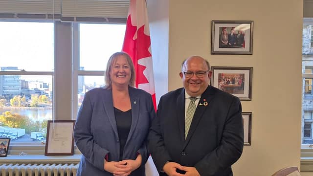 A woman in a gray blazer and Mayor Berry Vrbanovic in a dark suit smile side by side in an office beside a Canadian flag.