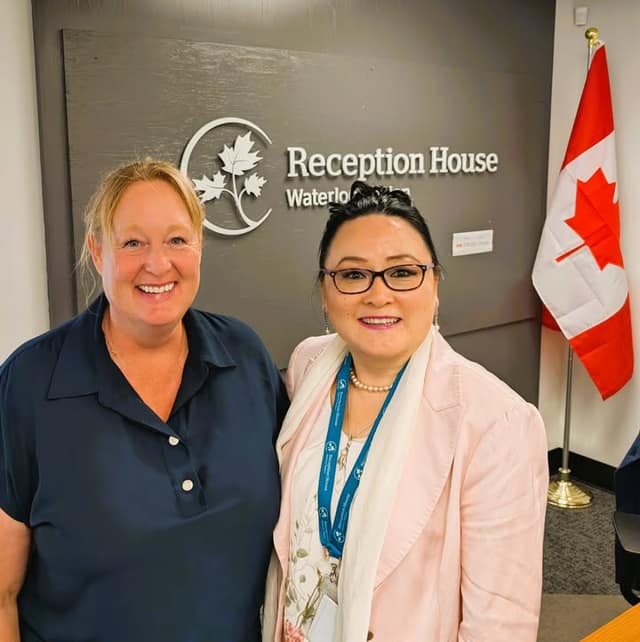 Two smiling women pose in front of a wall sign reading Reception House Waterloo Region, beside a Canadian flag.