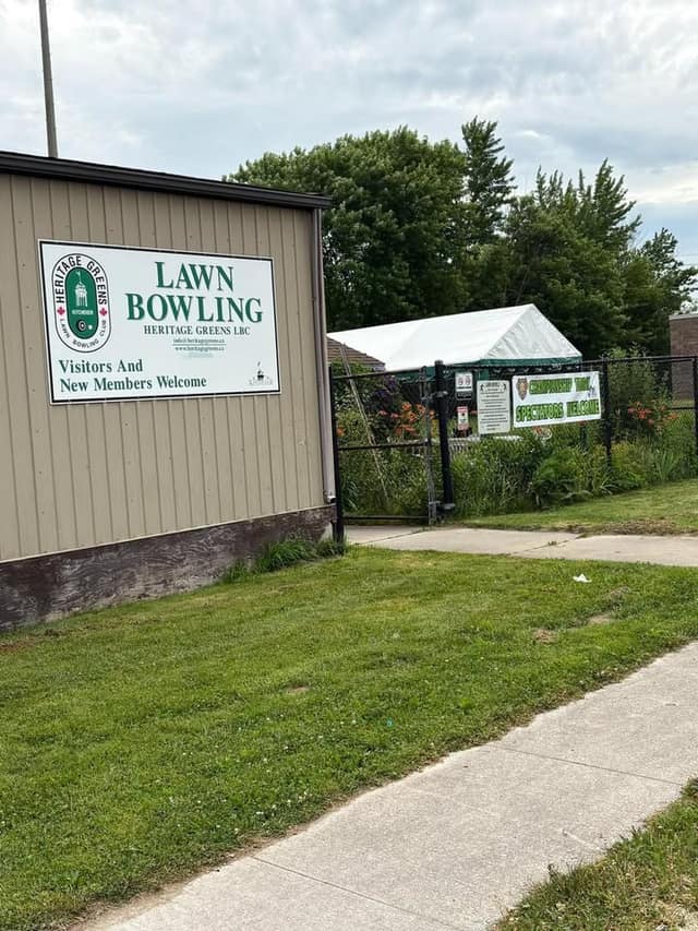 A sign on a tan building reads Lawn Bowling, Heritage Greens LBC, Visitors And New Members Welcome; a fence banner welcomes spectators.