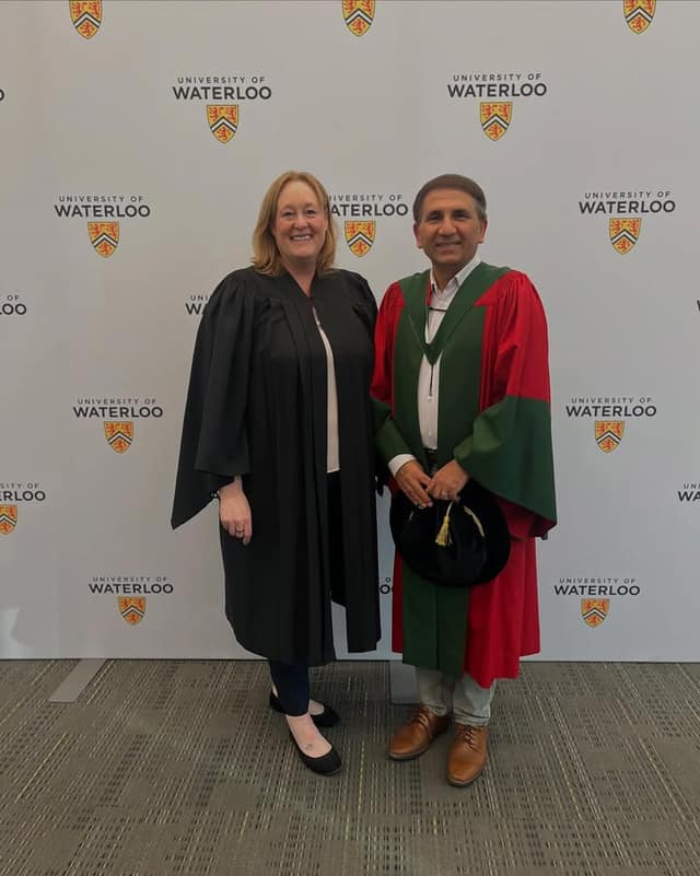 A smiling woman in a black gown stands beside a man in red and green academic robes holding a black cap, in front of a University of Waterloo backdrop.