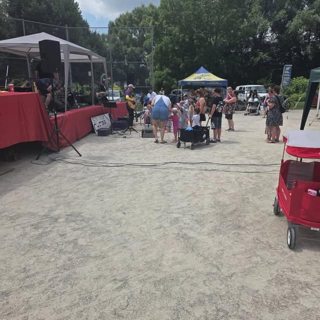 A musician plays guitar on a small stage beside speakers while families gather on a sandy area near tents and a red wagon.