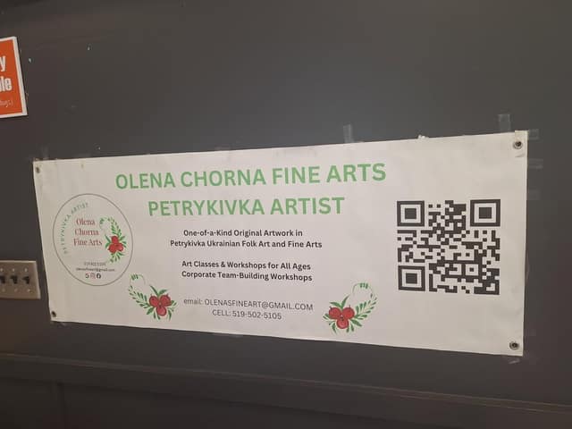A banner taped to a dark wall reads Olena Chorna Fine Arts, Petrykivka Artist, with art class details, contact info and a QR code.