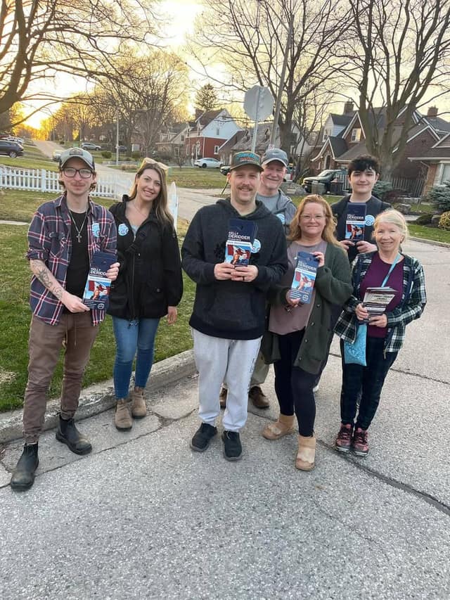 Six volunteers pose on a residential street holding Kelly DeRidder campaign door hangers, with houses and bare trees behind them.