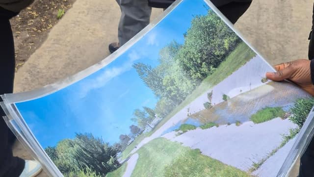 A hand holds a laminated printout showing a photo of a flooded section of paved path beside grass and trees under a blue sky.
