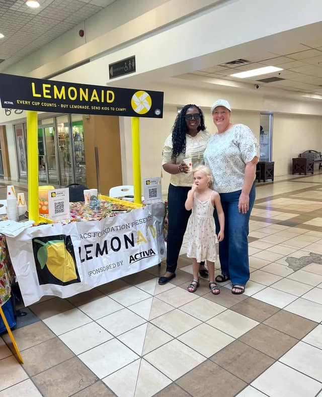 Two women and a young girl in a white dress stand beside the LemonAID stand in a mall, with a QR code sign on the table.