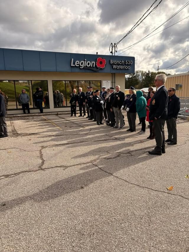 Veterans in blazers and berets and other attendees stand in a parking lot outside the Legion Branch 530 Waterloo building under a cloudy sky.