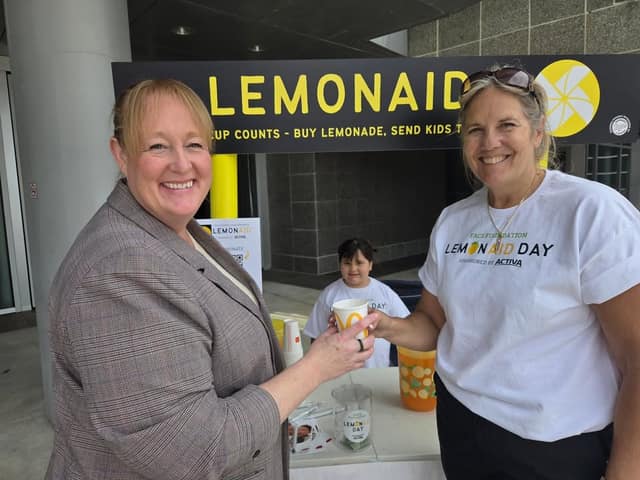 A smiling woman in a gray blazer takes a cup of lemonade from a smiling woman in a LemonAID Day shirt at a stand with a child behind.