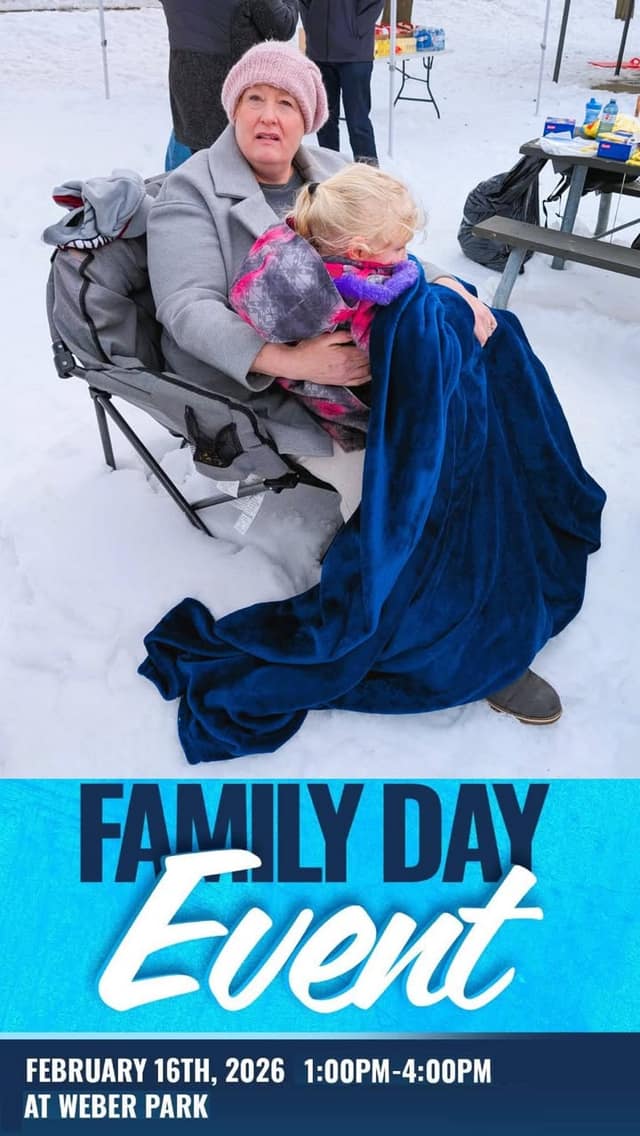 A woman in a pink toque and gray coat sits in a camp chair in the snow, holding a child wrapped in a blue blanket.
