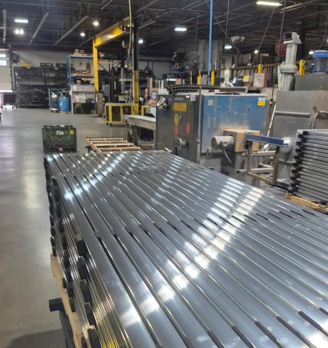 Stacked shiny metal profiles on a wooden pallet in the foreground of a factory floor with machinery, shelving and a green bin behind.