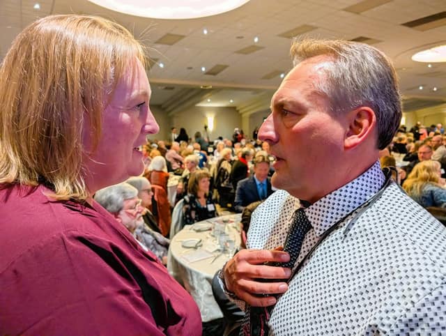 A woman in burgundy and a man in a dotted shirt and tie stand face to face in conversation, with a crowded banquet hall behind.