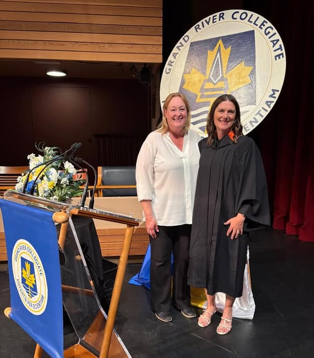Two smiling women stand on a stage beside a podium; one wears a black graduation gown, in front of a Grand River Collegiate sign.