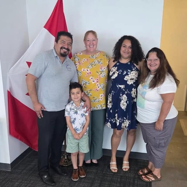 Four adults and a young boy in a floral shirt smile for a group photo in front of a Canadian flag in an office.