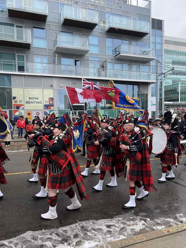A pipe band in red tartan kilts and white spats marches down a street, with Canadian and Ontario flags and a drummer behind.