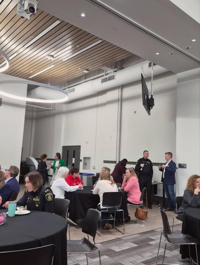 Attendees sit and stand in small groups around black-clothed round tables in a event room, including two uniformed police officers.