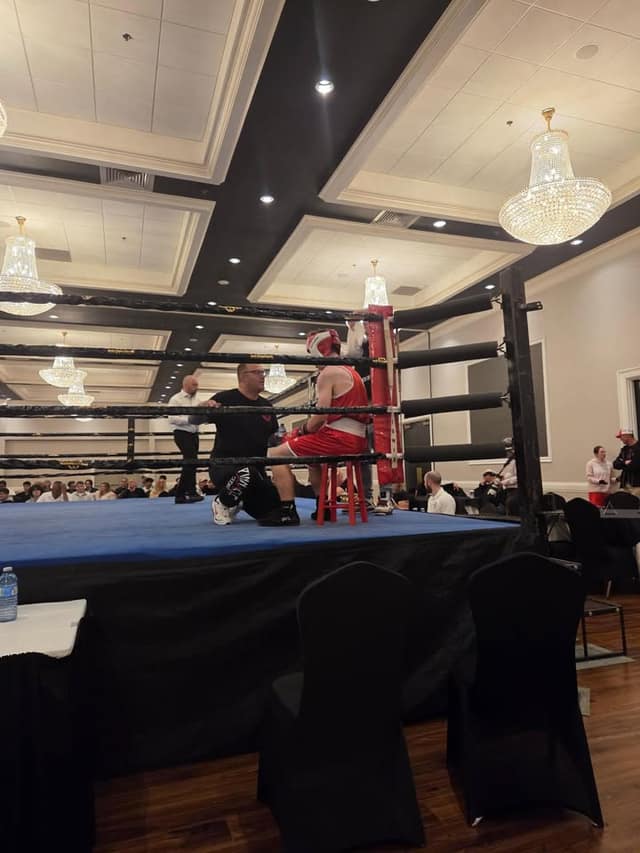 A boxer in red gear sits on a stool in a corner of the ring as a coach in black kneels beside him.