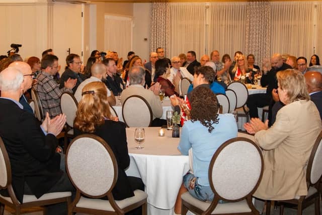 Guests seated at round tables with white cloths applaud in a banquet room with draped curtains and small Canadian flags.
