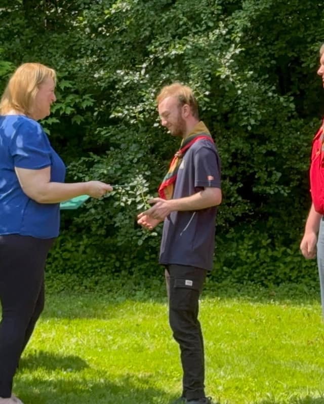 A woman in a blue top hands a green folder to a young man in a navy Scouts shirt and neckerchief on a grassy area.