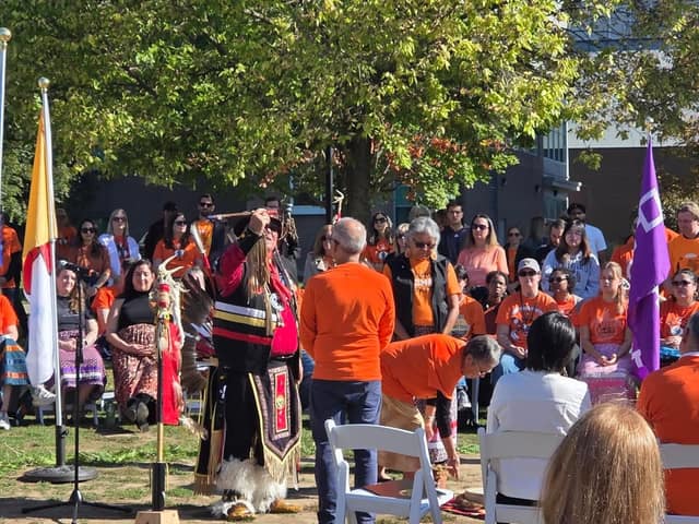 A regalia-clad man raises an instrument skyward at an outdoor ceremony as a seated crowd in orange shirts watches under a tree.