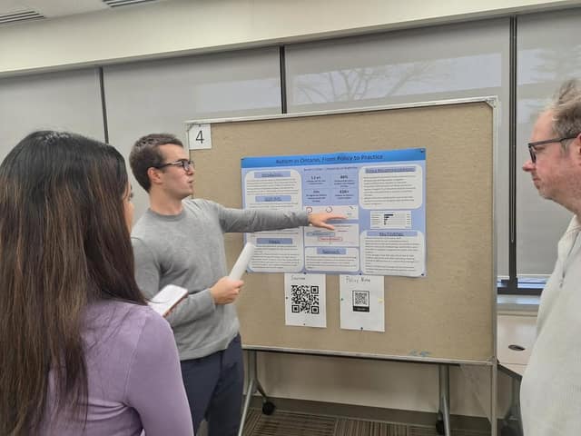 A student in glasses points to a research poster titled "Autism in Ontario, From Policy to Practice" while two people look on.