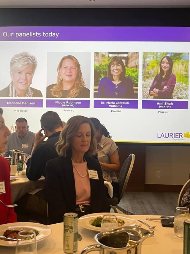 A projected slide titled "Our panelists today" shows four portraits, with luncheon guests seated at tables below the screen.