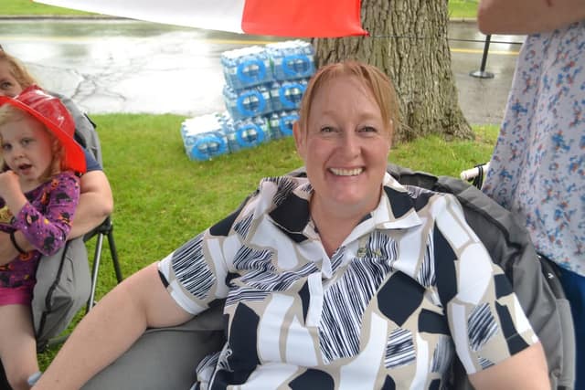 A smiling woman in a black, white and tan patterned shirt sits in a folding chair on grass, with stacked water bottles and a child behind her.