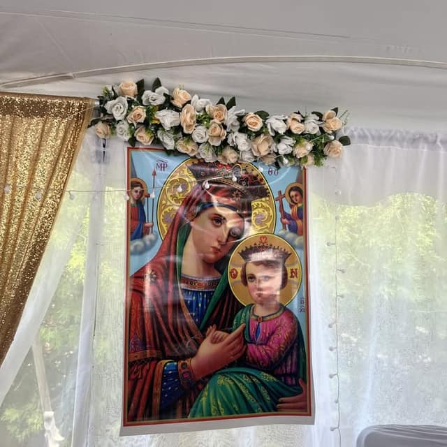 A religious icon of Mary holding the child Jesus hangs under a white tent, topped with cream and white roses and beside a gold sequin curtain.