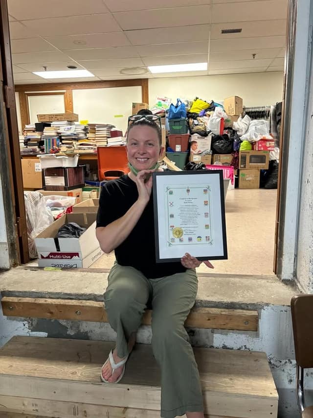 A smiling woman sits on a wooden step holding a framed certificate, with boxes and donated goods piled in the room behind her.