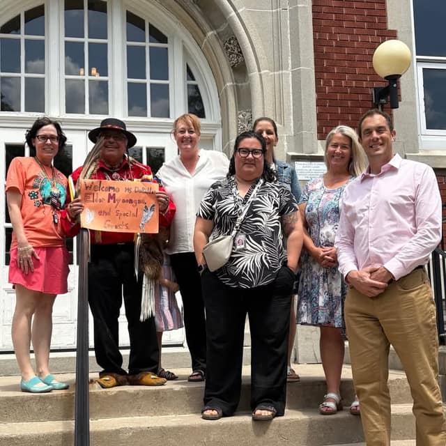Six adults stand on school steps; Elder Myeengun holds an orange sign reading "Welcome" to the Elder and special guests.